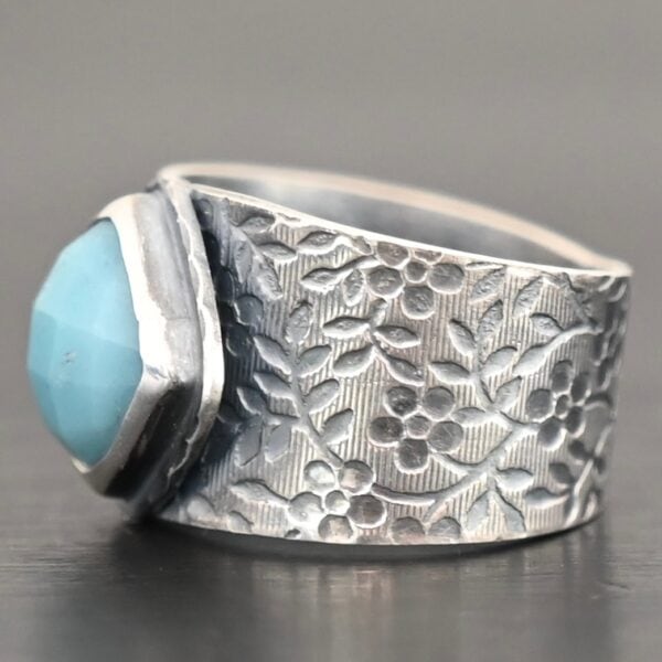 Photo of the "Naia" Ring. There is a medium sized, diamond shaped aquamarine in the center of the ring. The ring band is wide, oxidized lightly, sterling silver, with flower designs etched all the way around the entire band. Ring is sitting against a dark background.