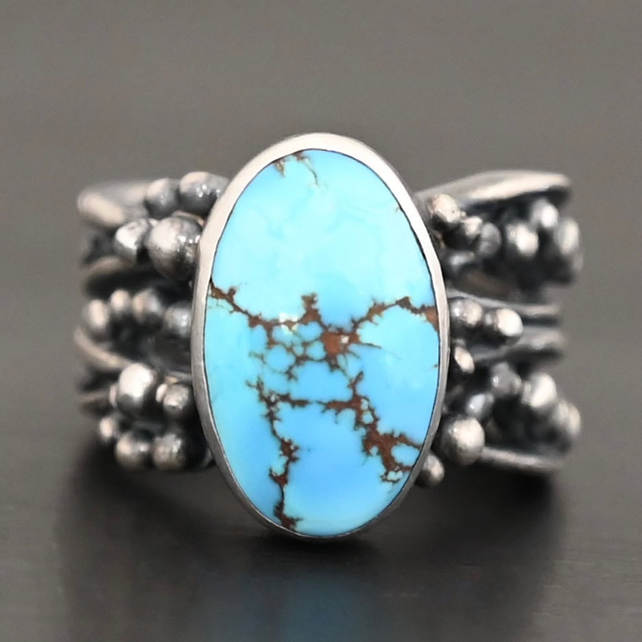 Eira Ring Photo of the "Eira" Ring. Made out of sterling silver and turquoise. There is a medium sized, oval turquoise set vertically in the center. The rest of the ring band is made out of multiple lines of silver that are wavy. They intercept at random areas and leave small gaps in the ring band. There are multiple balls of silver around the rest of the ring band.