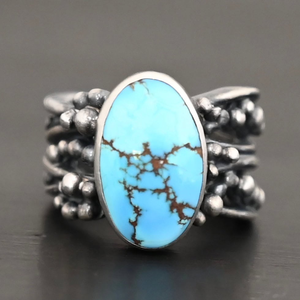 Eira Ring Photo of the "Eira" Ring. Made out of sterling silver and turquoise. There is a medium sized, oval turquoise set vertically in the center. The rest of the ring band is made out of multiple lines of silver that are wavy. They intercept at random areas and leave small gaps in the ring band. There are multiple balls of silver around the rest of the ring band.
