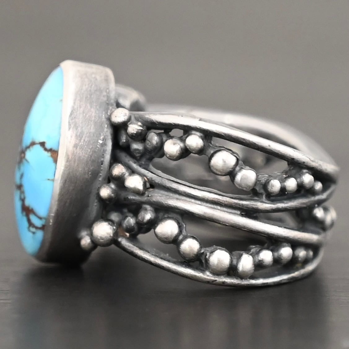 Eira Ring Photo of the "Eira" Ring. Made out of sterling silver and turquoise. There is a medium sized, oval turquoise set vertically in the center. The rest of the ring band is made out of multiple lines of silver that are wavy. They intercept at random areas and leave small gaps in the ring band. There are multiple balls of silver around the rest of the ring band.