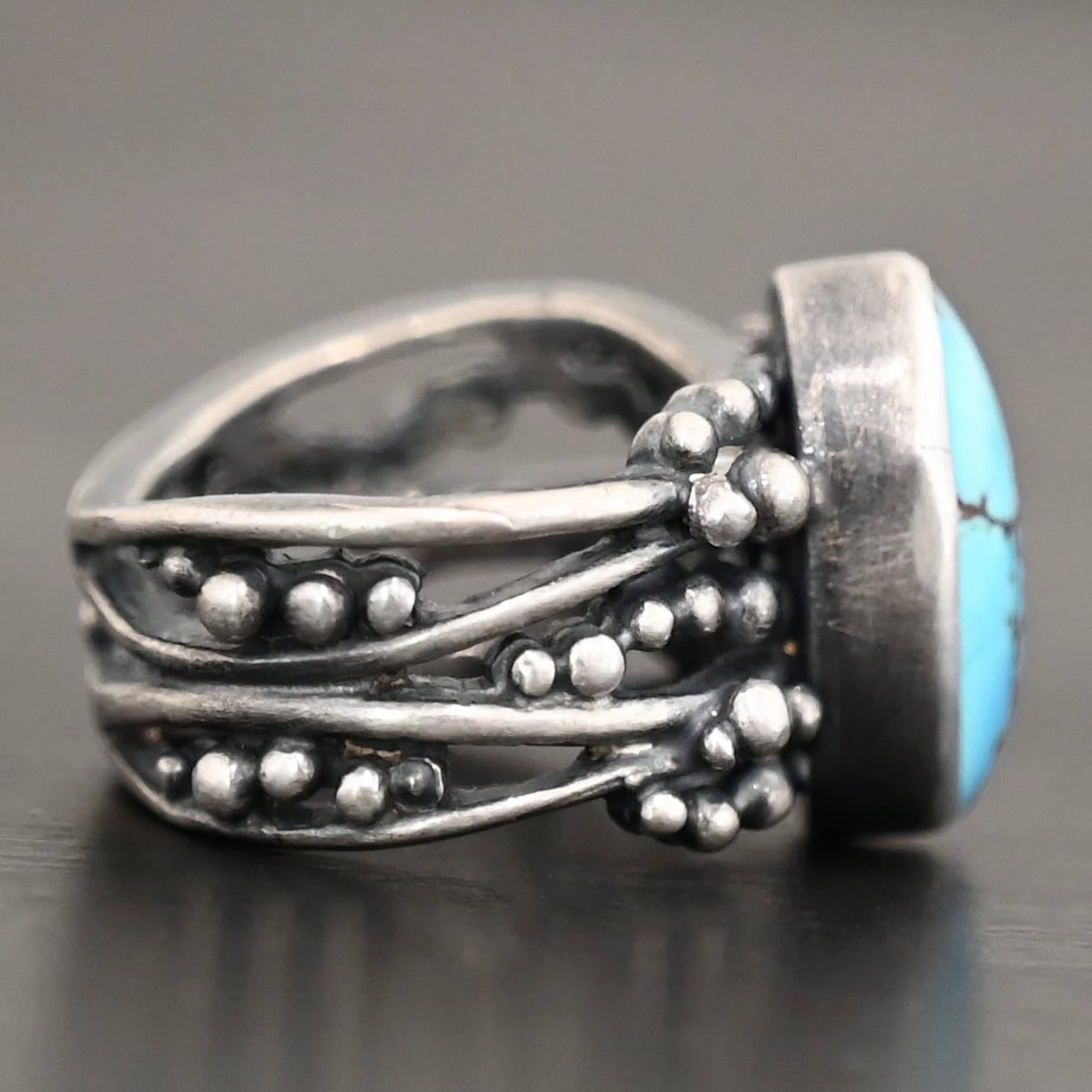 Eira Ring Photo of the "Eira" Ring. Made out of sterling silver and turquoise. There is a medium sized, oval turquoise set vertically in the center. The rest of the ring band is made out of multiple lines of silver that are wavy. They intercept at random areas and leave small gaps in the ring band. There are multiple balls of silver around the rest of the ring band.