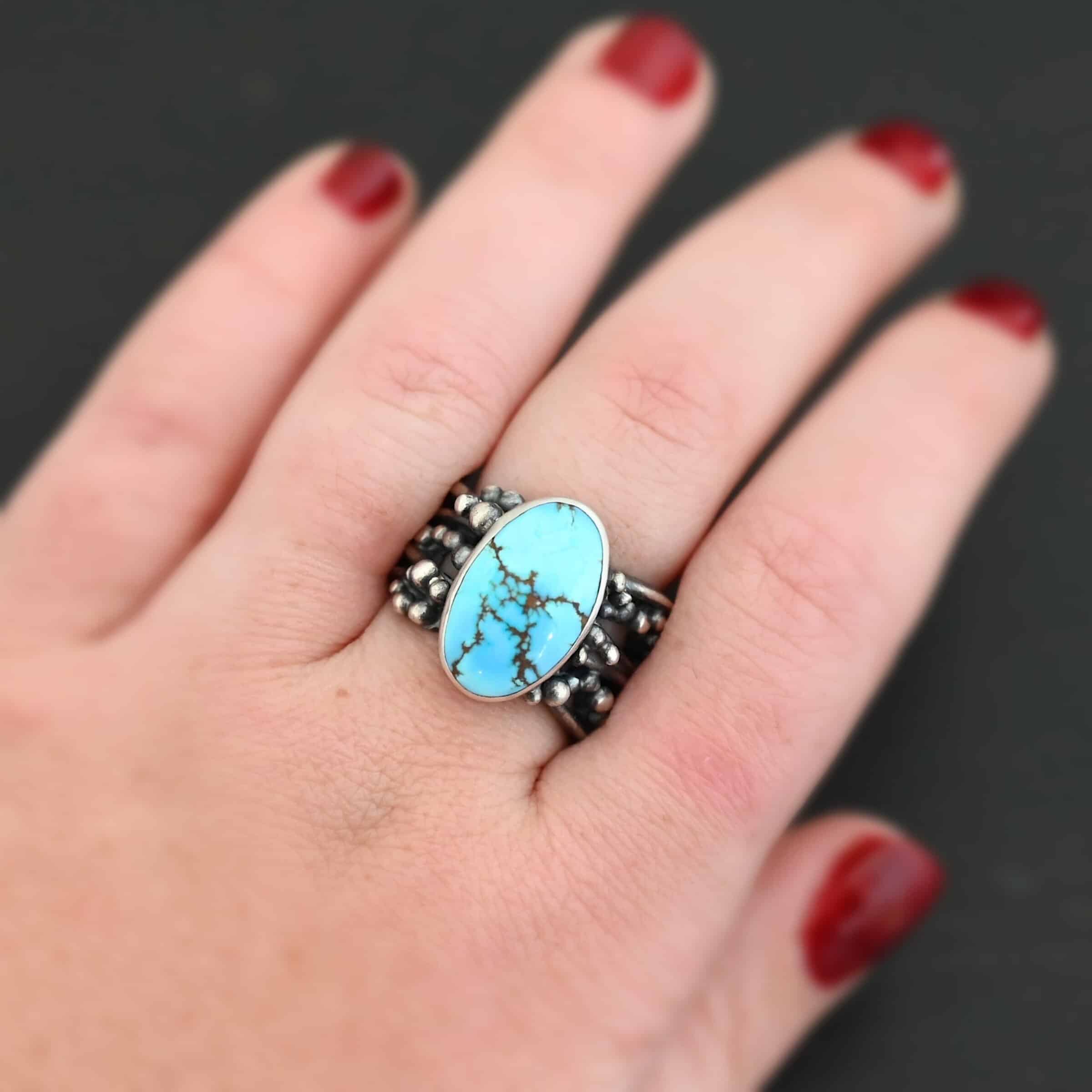 Eira Ring Photo of the "Eira" Ring. Made out of sterling silver and turquoise. There is a medium sized, oval turquoise set vertically in the center. The rest of the ring band is made out of multiple lines of silver that are wavy. They intercept at random areas and leave small gaps in the ring band. There are multiple balls of silver around the rest of the ring band. Ring is worn by a model to demonstrate size.
