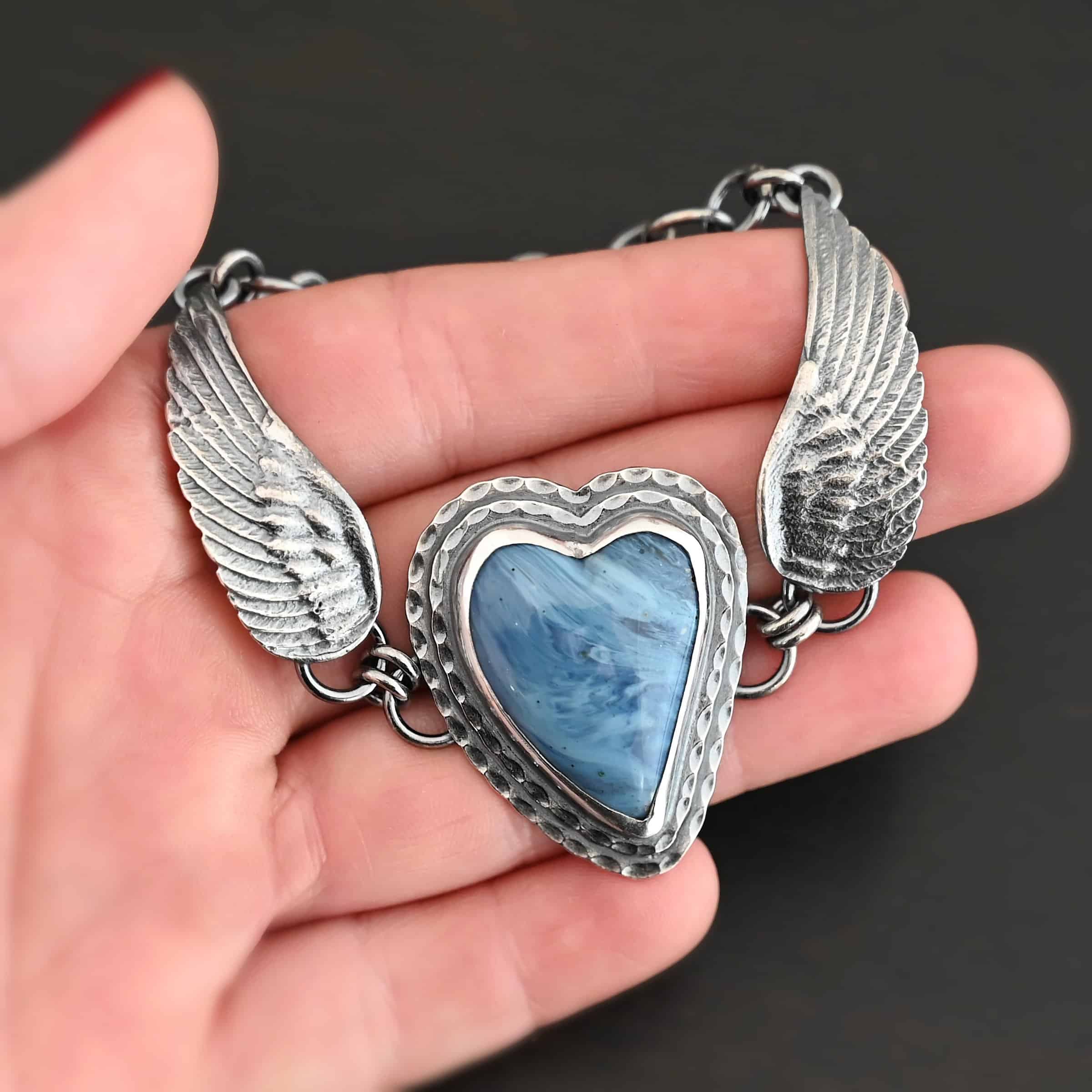 Waves Of Love Bracelet "Waves Of Love" Bracelet. The bracelet is a Leland Blue heart with medium sized angel wings on each side. The angel wings curve lightly so they can fit around the wrist. There is two layers of hammered sterling silver around the heart. Bracelet has a silver lobster claw clasp. Bracelet is shown up close in the fingertips of a hand.