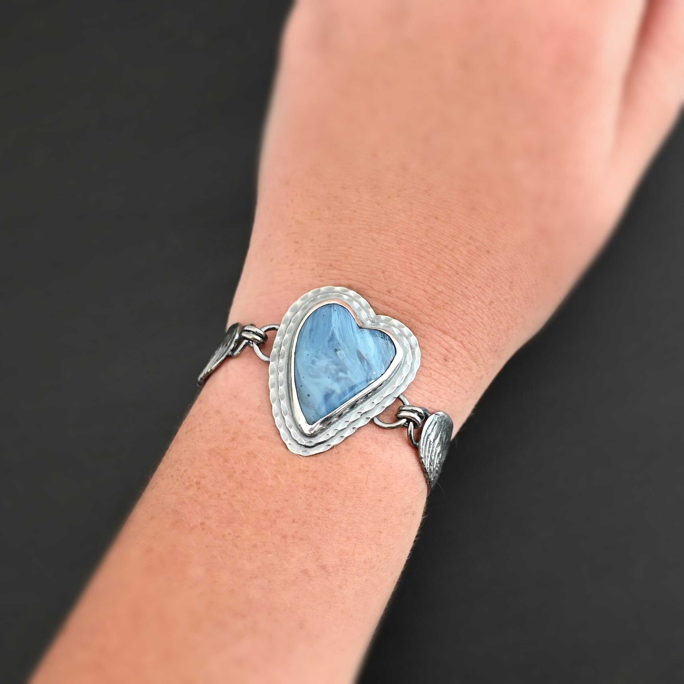 Waves Of Love Bracelet "Waves Of Love" Bracelet. The bracelet is a Leland Blue heart with medium sized angel wings on each side. The angel wings curve lightly so they can fit around the wrist. There is two layers of hammered sterling silver around the heart. Bracelet has a silver lobster claw clasp. Bracelet is shown up close by a model wearing it on their wrist.