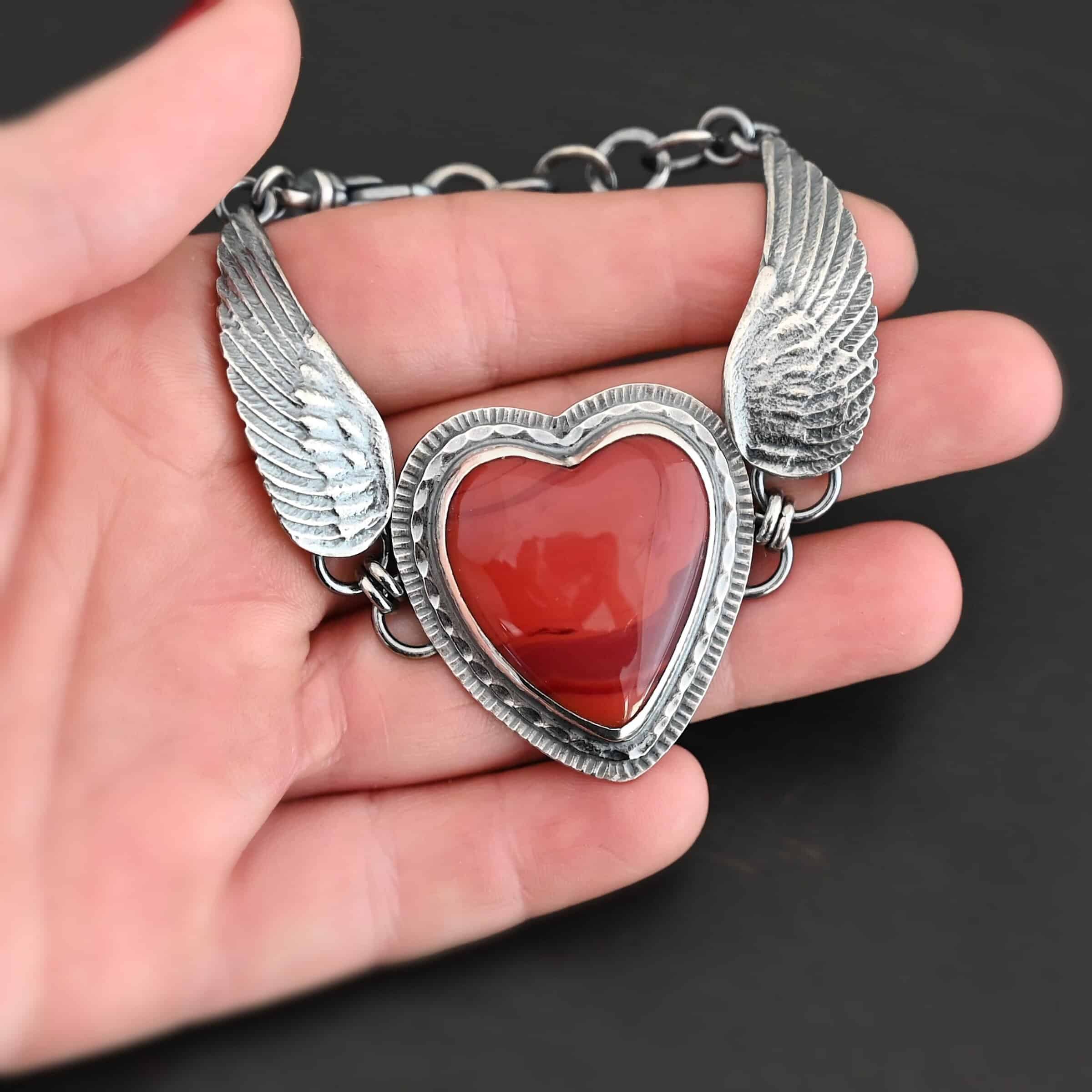 "Cerise" Bracelet. The bracelet is a rosarita heart with medium sized angel wings on each side. The angel wings curve lightly so they can fit around the wrist. There is two layers of hammered sterling silver around the heart. Bracelet has a silver lobster claw clasp. Bracelet is held in the fingertips of a hand.