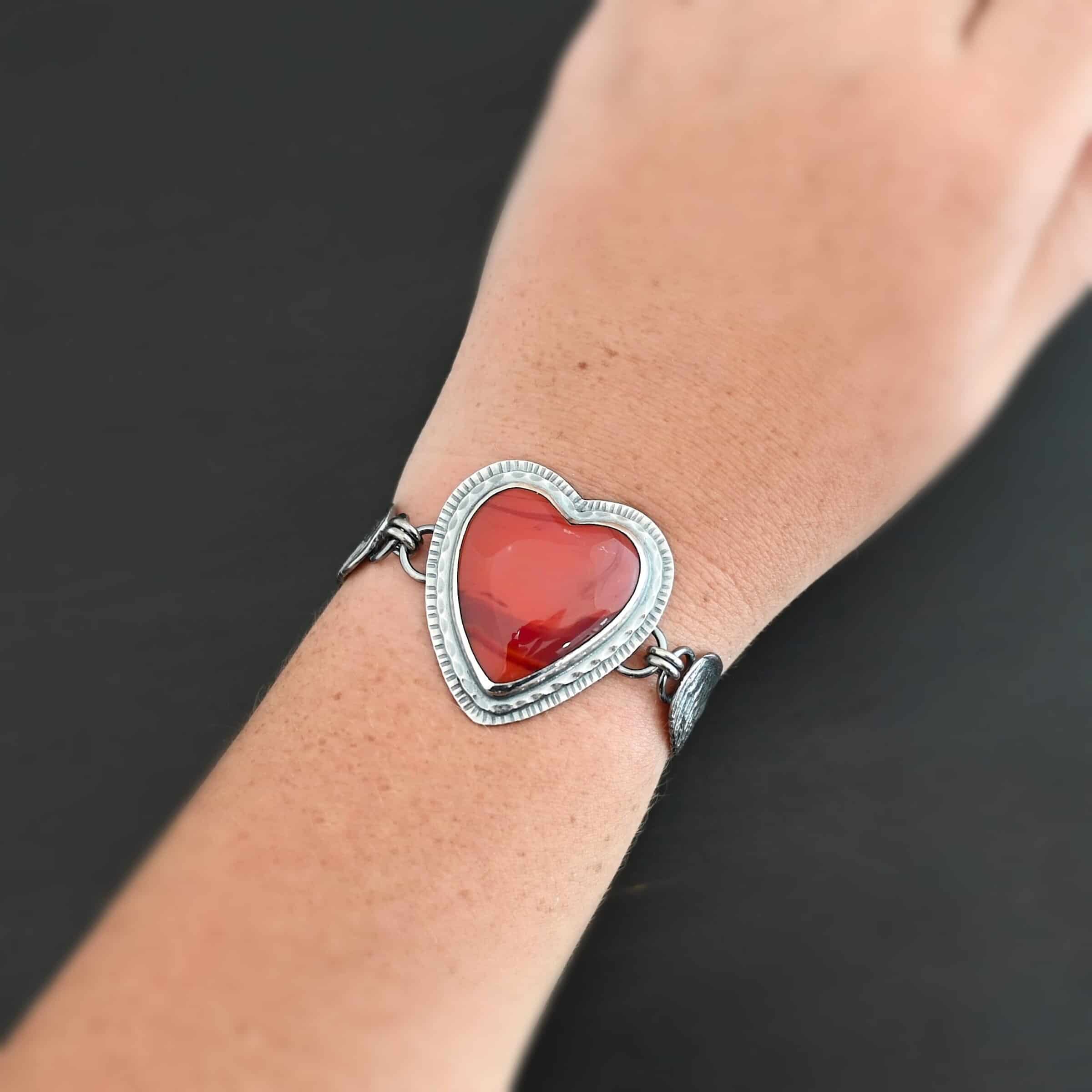 "Cerise" Bracelet. The bracelet is a rosarita heart with medium sized angel wings on each side. The angel wings curve lightly so they can fit around the wrist. There is two layers of hammered sterling silver around the heart. Bracelet has a silver lobster claw clasp. Bracelet is shown up close by a model on their wrist.