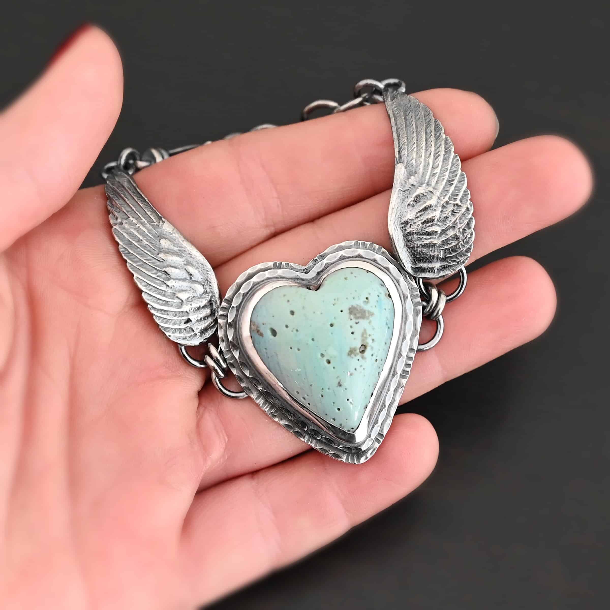 Vanessa Bracelet "Vanessa" Bracelet. The bracelet is a Leland Blue heart with medium sized angel wings on each side. The angel wings curve lightly so they can fit around the wrist. There is two layers of hammered sterling silver around the heart. Bracelet has a silver lobster claw clasp. Bracelet is held in the fingertips of a hand.