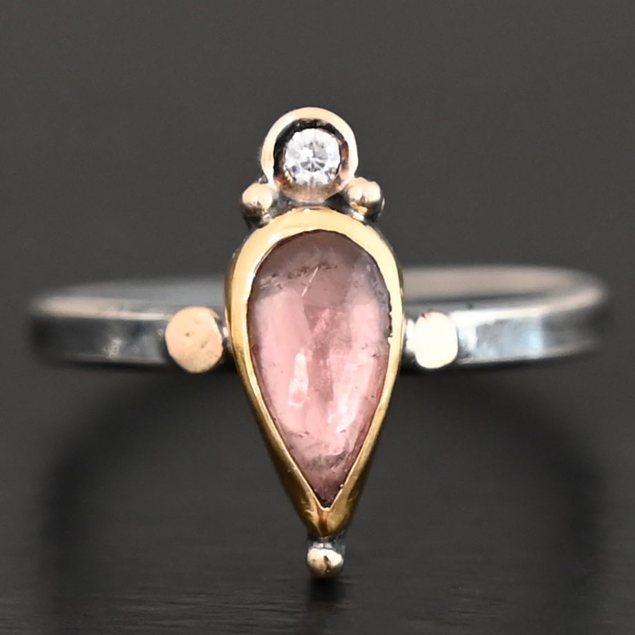 "Elle" Ring. There is a pear shaped pink tourmaline pointing downwards. It is set in gold. There is a small diamond above it, set in gold, and with a gold ball on each side of it. There is a gold ball on the bottom of the point of the tourmaline. There are two dots of gold, one on each side of the tourmaline, set on the silver ring band.