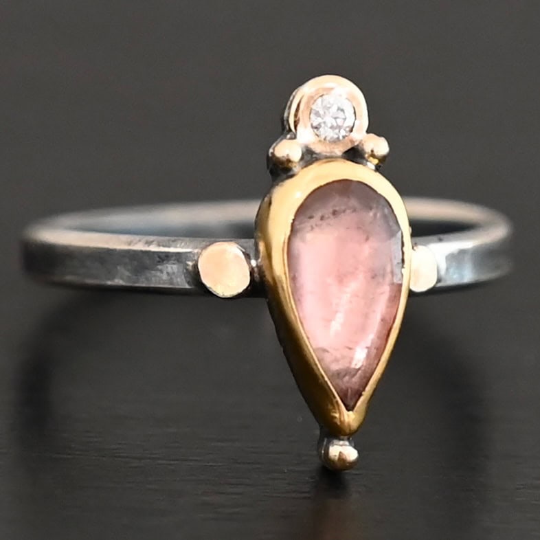 "Elle" Ring. There is a pear shaped pink tourmaline pointing downwards. It is set in gold. There is a small diamond above it, set in gold, and with a gold ball on each side of it. There is a gold ball on the bottom of the point of the tourmaline. There are two dots of gold, one on each side of the tourmaline, set on the silver ring band.