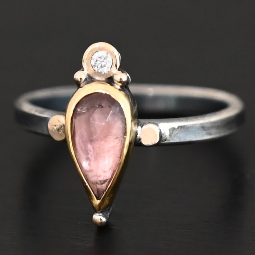 "Elle" Ring. There is a pear shaped pink tourmaline pointing downwards. It is set in gold. There is a small diamond above it, set in gold, and with a gold ball on each side of it. There is a gold ball on the bottom of the point of the tourmaline. There are two dots of gold, one on each side of the tourmaline, set on the silver ring band.