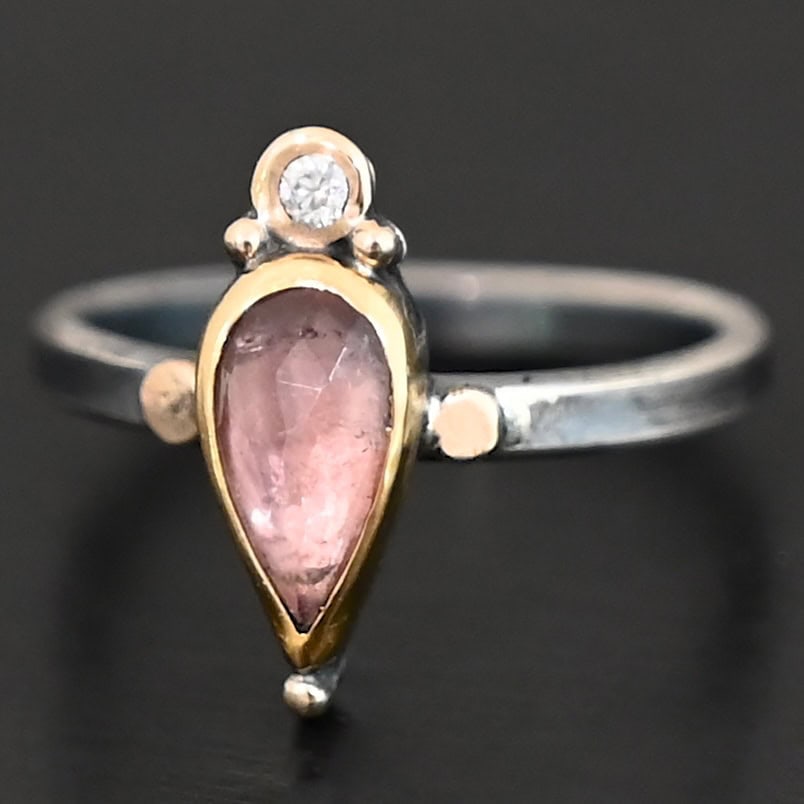 "Elle" Ring. There is a pear shaped pink tourmaline pointing downwards. It is set in gold. There is a small diamond above it, set in gold, and with a gold ball on each side of it. There is a gold ball on the bottom of the point of the tourmaline. There are two dots of gold, one on each side of the tourmaline, set on the silver ring band.