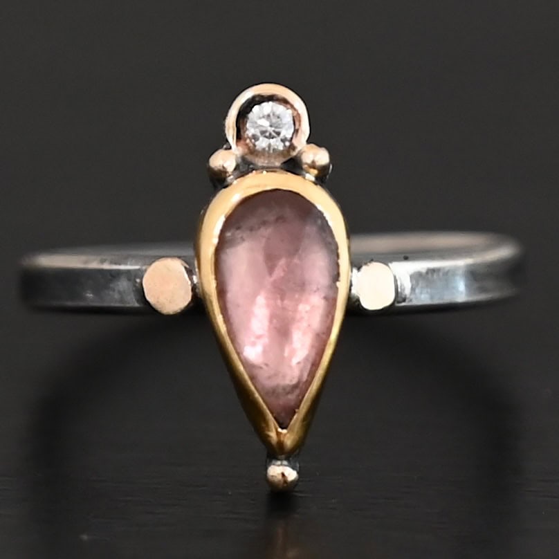 "Elle" Ring. There is a pear shaped pink tourmaline pointing downwards. It is set in gold. There is a small diamond above it, set in gold, and with a gold ball on each side of it. There is a gold ball on the bottom of the point of the tourmaline. There are two dots of gold, one on each side of the tourmaline, set on the silver ring band.