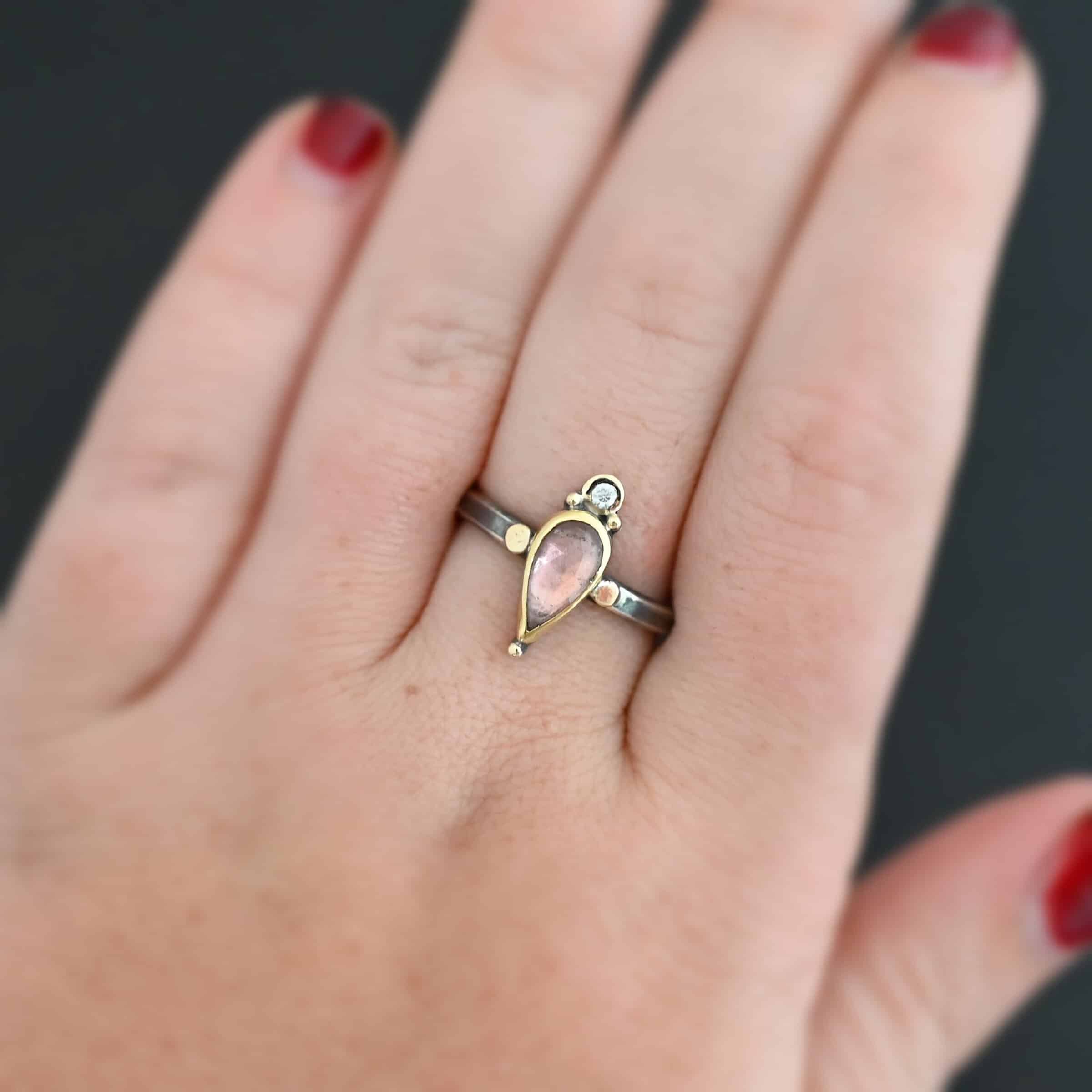 "Elle" Ring. There is a pear shaped pink tourmaline pointing downwards. It is set in gold. There is a small diamond above it, set in gold, and with a gold ball on each side of it. There is a gold ball on the bottom of the point of the tourmaline. There are two dots of gold, one on each side of the tourmaline, set on the silver ring band. Ring is shown up close by a model.