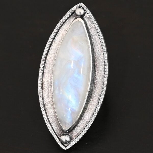 "Artemis Ring" The ring is large, marquise, vertical moonstone. There is a ball of silver on the top and bottom of it. There is textured silver around it, and a thin layer around that which is hammered.