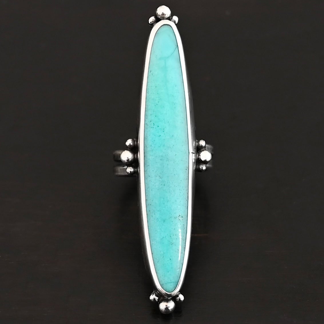 "Abilene" Ring. It is a long, oval, vertical turquoise. There are four groups of three balls of silver around the ring. Three are on the top, three on the bottom, and on the left and right side.