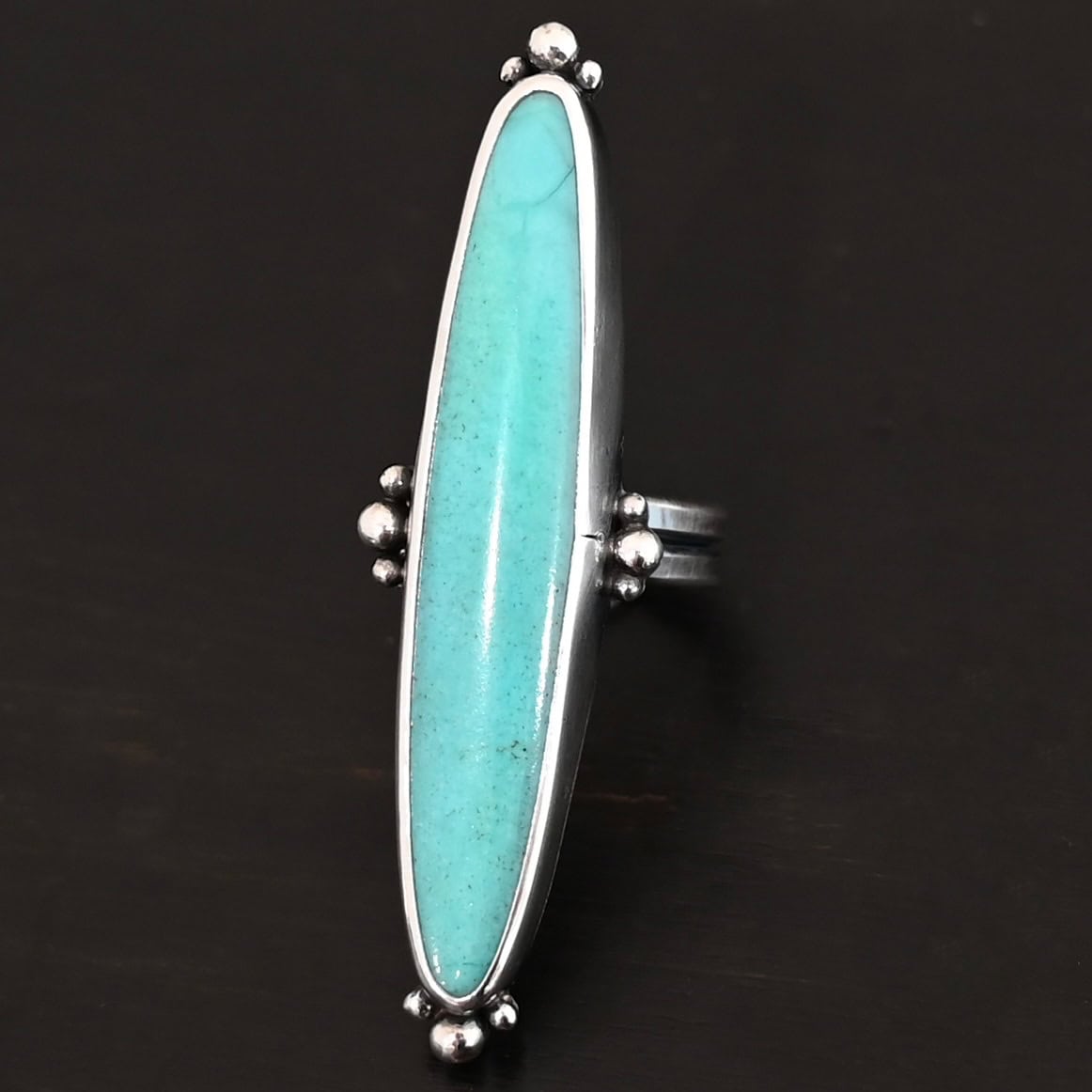 "Abilene" Ring. It is a long, oval, vertical turquoise. There are four groups of three balls of silver around the ring. Three are on the top, three on the bottom, and on the left and right side.