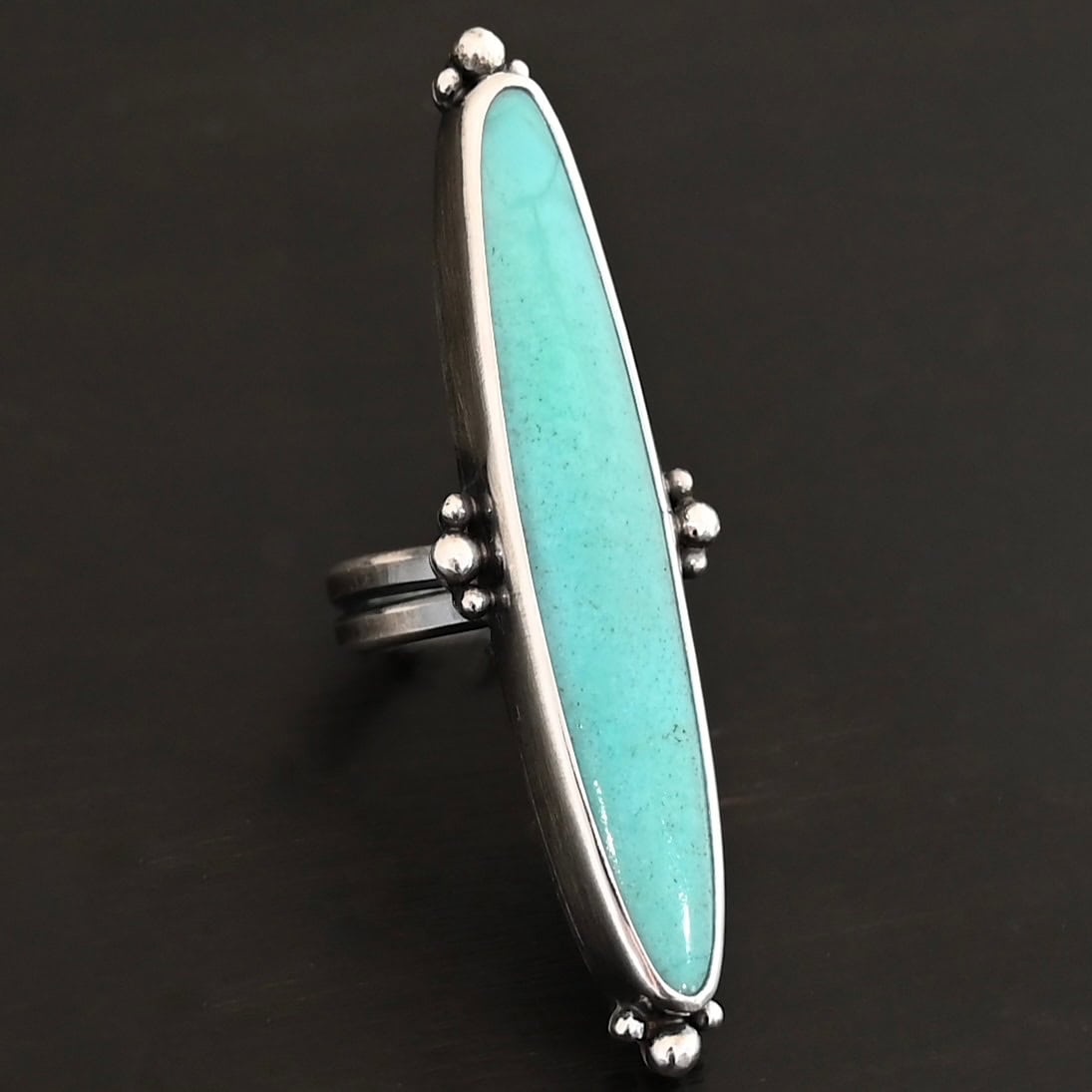 "Abilene" Ring. It is a long, oval, vertical turquoise. There are four groups of three balls of silver around the ring. Three are on the top, three on the bottom, and on the left and right side.