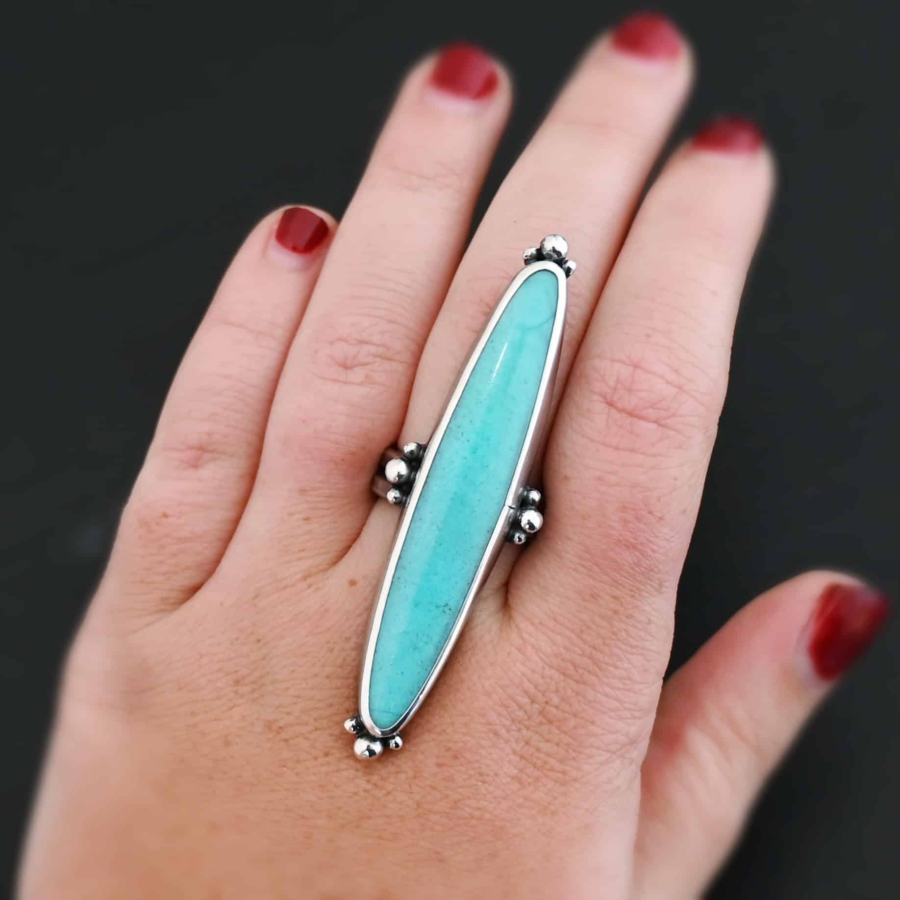 "Abilene" Ring. It is a long, oval, vertical turquoise. There are four groups of three balls of silver around the ring. Three are on the top, three on the bottom, and on the left and right side. Ring is shown up close by a model.
