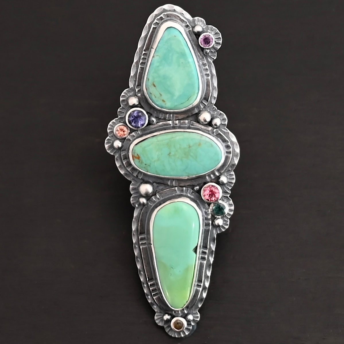 "Western Flowers" Ring. There are three oval turquoise. The one on the top and bottom are vertical, and the one in between is horizontal. They are all a larger size. There are six other, smaller circular stones scattered around the turquoise. There are two layers of hammered silver around all of the stones.
