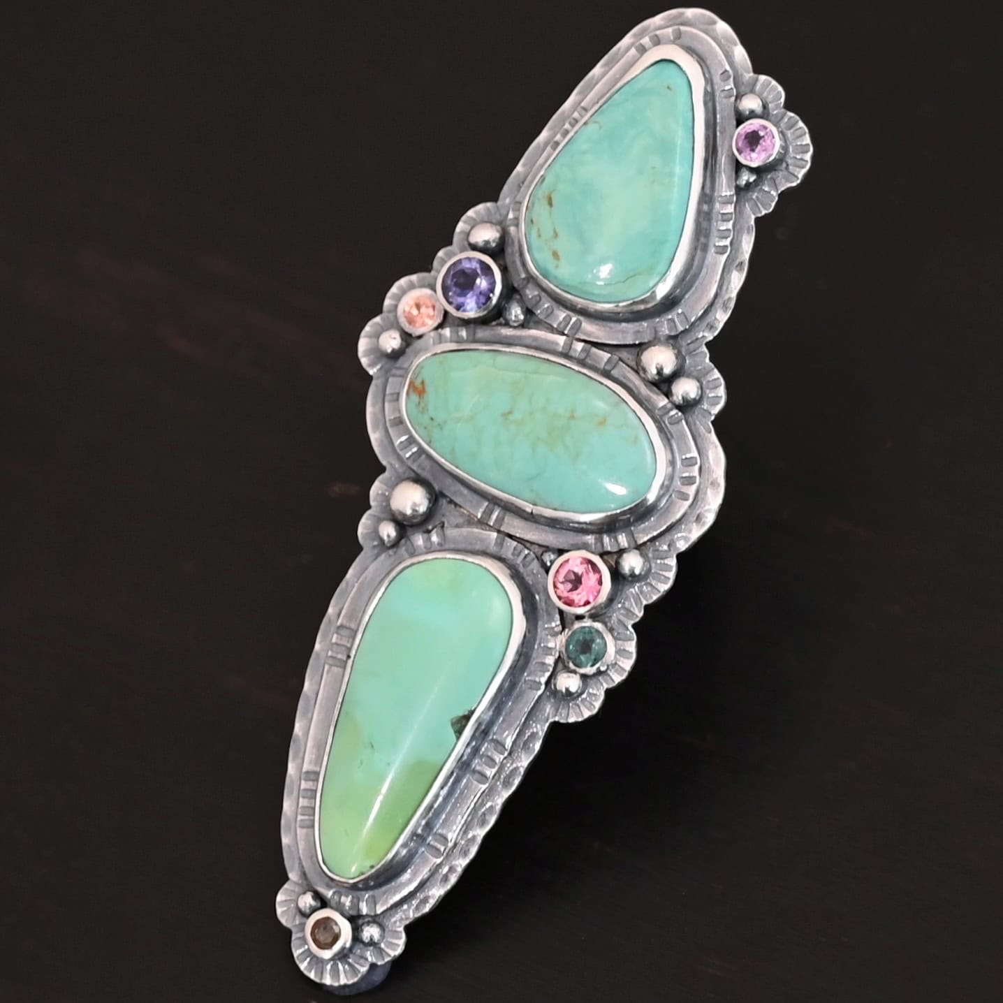 "Western Flowers" Ring. There are three oval turquoise. The one on the top and bottom are vertical, and the one in between is horizontal. They are all a larger size. There are six other, smaller circular stones scattered around the turquoise. There are two layers of hammered silver around all of the stones.