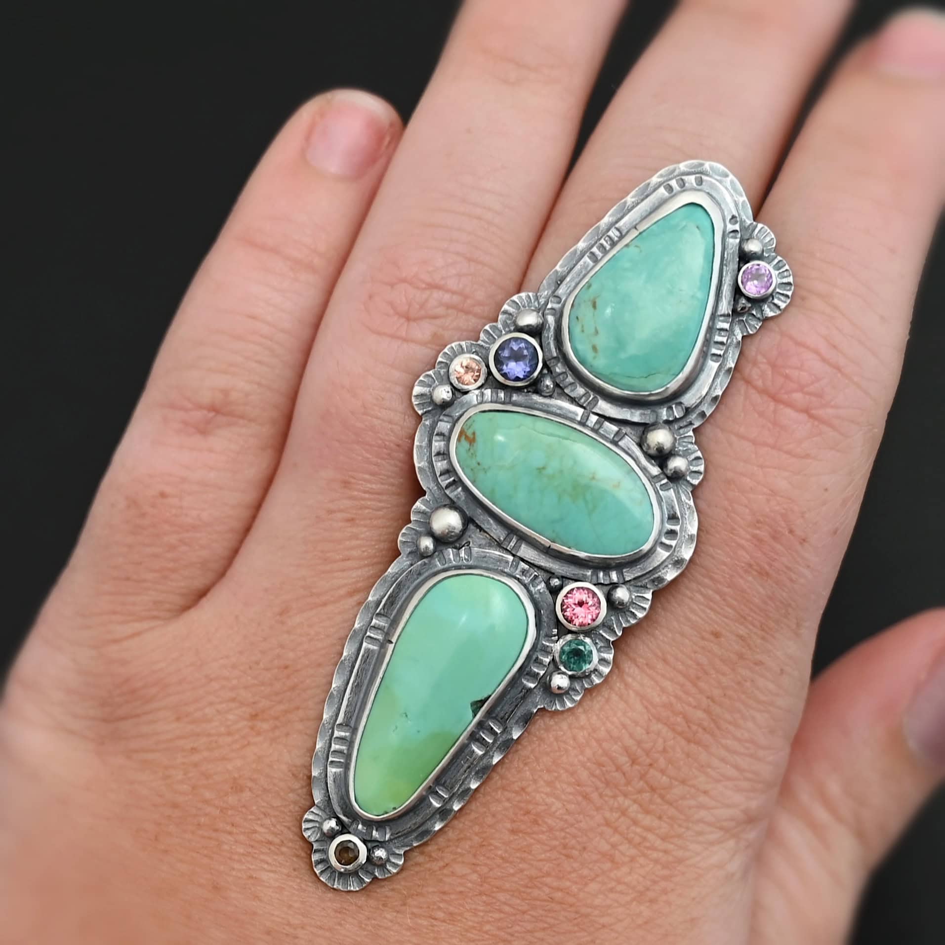 "Western Flowers" Ring. There are three oval turquoise. The one on the top and bottom are vertical, and the one in between is horizontal. They are all a larger size. There are six other, smaller circular stones scattered around the turquoise. There are two layers of hammered silver around all of the stones. Ring is shown on a model's hand up close.