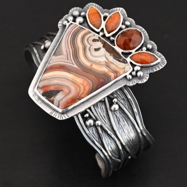 "Something In The Orange" Cuff. Sterling Silver, Teepee Canyon Agate, and Orange Kyanite. There is a large, wide, trapezoid agate on the bottom with four orange kyanites going across it on the top. Three are marquise shaped and one is a circular shape. The cuff band is very wide, and has lines of silver that are wavy and go across it. There is silver ball detail around the rest of the cuff.