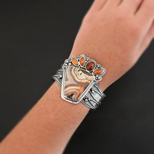 "Something In The Orange" Cuff. Sterling Silver, Teepee Canyon Agate, and Orange Kyanite. There is a large, wide, trapezoid agate on the bottom with four orange kyanites going across it on the top. Three are marquise shaped and one is a circular shape. The cuff band is very wide, and has lines of silver that are wavy and go across it. There is silver ball detail around the rest of the cuff. Cuff is shown on a model up close.