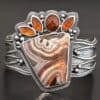 "Something In The Orange" Cuff. Sterling Silver, Teepee Canyon Agate, and Orange Kyanite. There is a large, wide, trapezoid agate on the bottom with four orange kyanites going across it on the top. Three are marquise shaped and one is a circular shape. The cuff band is very wide, and has lines of silver that are wavy and go across it. There is silver ball detail around the rest of the cuff.