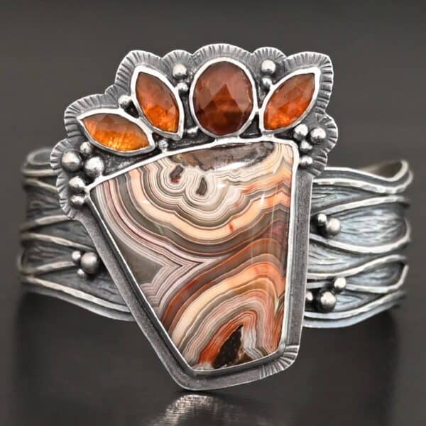 "Something In The Orange" Cuff. Sterling Silver, Teepee Canyon Agate, and Orange Kyanite. There is a large, wide, trapezoid agate on the bottom with four orange kyanites going across it on the top. Three are marquise shaped and one is a circular shape. The cuff band is very wide, and has lines of silver that are wavy and go across it. There is silver ball detail around the rest of the cuff.