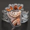 "Something In The Orange" Cuff. Sterling Silver, Teepee Canyon Agate, and Orange Kyanite. There is a large, wide, trapezoid agate on the bottom with four orange kyanites going across it on the top. Three are marquise shaped and one is a circular shape. The cuff band is very wide, and has lines of silver that are wavy and go across it. There is silver ball detail around the rest of the cuff.