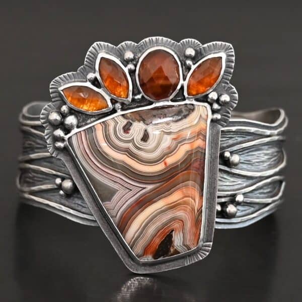 "Something In The Orange" Cuff. Sterling Silver, Teepee Canyon Agate, and Orange Kyanite. There is a large, wide, trapezoid agate on the bottom with four orange kyanites going across it on the top. Three are marquise shaped and one is a circular shape. The cuff band is very wide, and has lines of silver that are wavy and go across it. There is silver ball detail around the rest of the cuff.