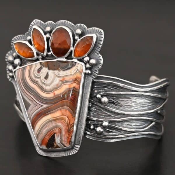 "Something In The Orange" Cuff. Sterling Silver, Teepee Canyon Agate, and Orange Kyanite. There is a large, wide, trapezoid agate on the bottom with four orange kyanites going across it on the top. Three are marquise shaped and one is a circular shape. The cuff band is very wide, and has lines of silver that are wavy and go across it. There is silver ball detail around the rest of the cuff.