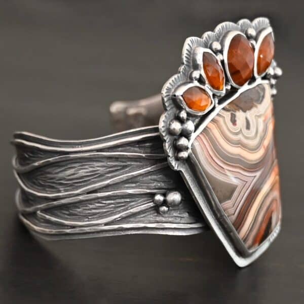 "Something In The Orange" Cuff. Sterling Silver, Teepee Canyon Agate, and Orange Kyanite. There is a large, wide, trapezoid agate on the bottom with four orange kyanites going across it on the top. Three are marquise shaped and one is a circular shape. The cuff band is very wide, and has lines of silver that are wavy and go across it. There is silver ball detail around the rest of the cuff.