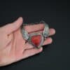 "Cerise" Bracelet. The bracelet is a rosarita heart with medium sized angel wings on each side. The angel wings curve lightly so they can fit around the wrist. There is two layers of hammered sterling silver around the heart. Bracelet has a silver lobster claw clasp. Bracelet is held in the fingertips of a hand.