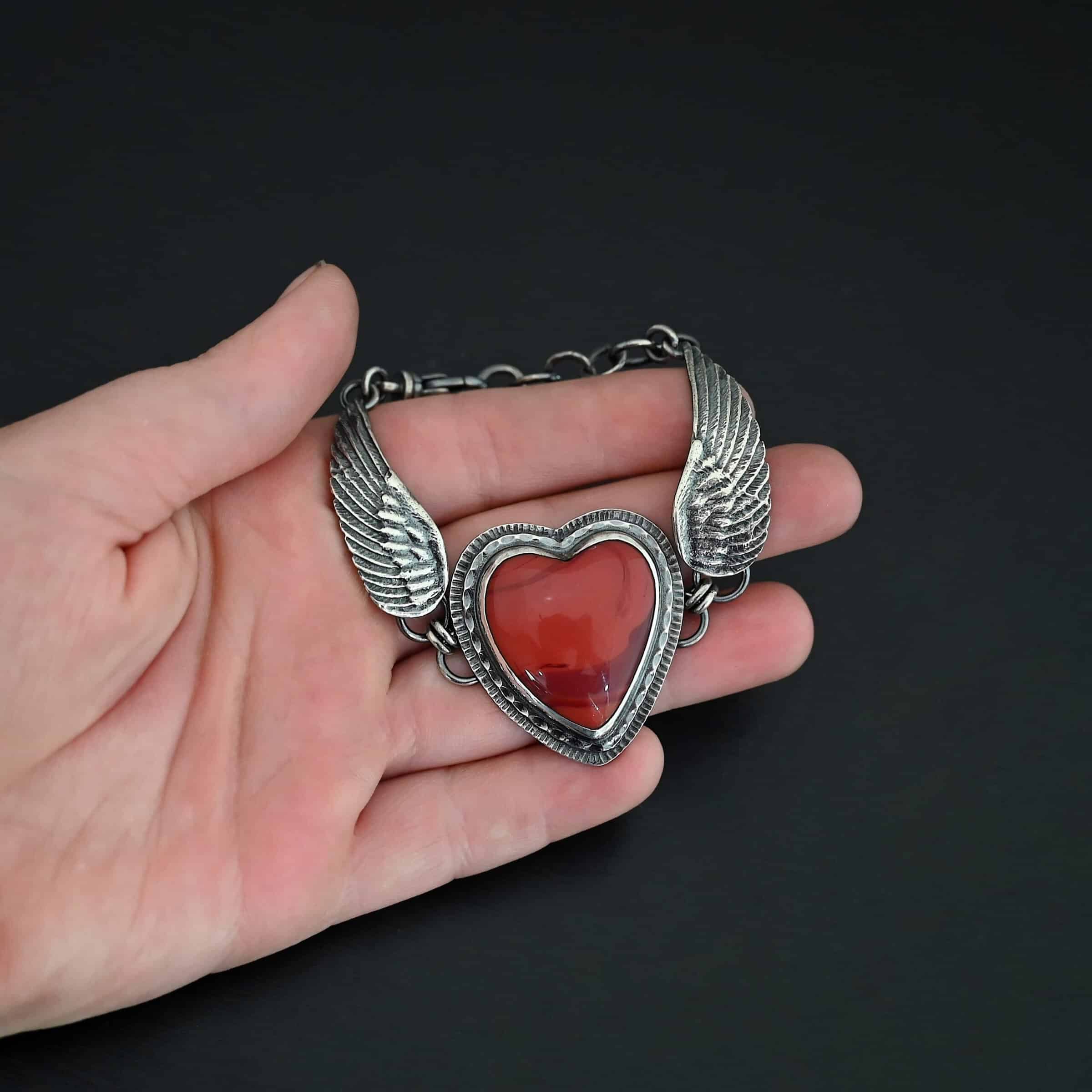 "Cerise" Bracelet. The bracelet is a rosarita heart with medium sized angel wings on each side. The angel wings curve lightly so they can fit around the wrist. There is two layers of hammered sterling silver around the heart. Bracelet has a silver lobster claw clasp. Bracelet is held in the fingertips of a hand.