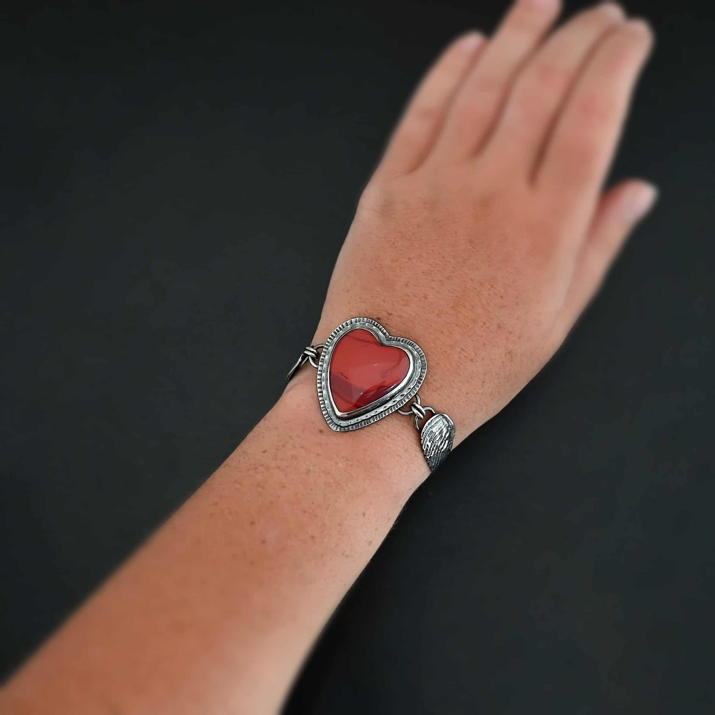 "Cerise" Bracelet. The bracelet is a rosarita heart with medium sized angel wings on each side. The angel wings curve lightly so they can fit around the wrist. There is two layers of hammered sterling silver around the heart. Bracelet has a silver lobster claw clasp. Bracelet is shown up close by a model on their wrist.