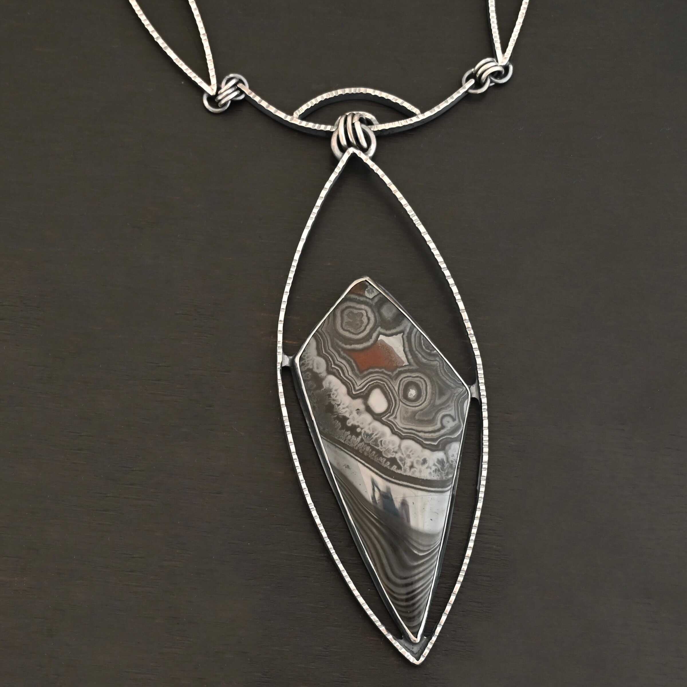 Zinnia Necklace New "Zinnia" Necklace. There is a large, diamond shaped black, grey, and red stone. There is a marquise shape of silver that goes around the stone, connected on each side to the stone. The chain of the necklace has two marquise shapes of silver, one on each side. The center is a small curve of silver from which the jump rings hang from that are attached to the marquise of silver.