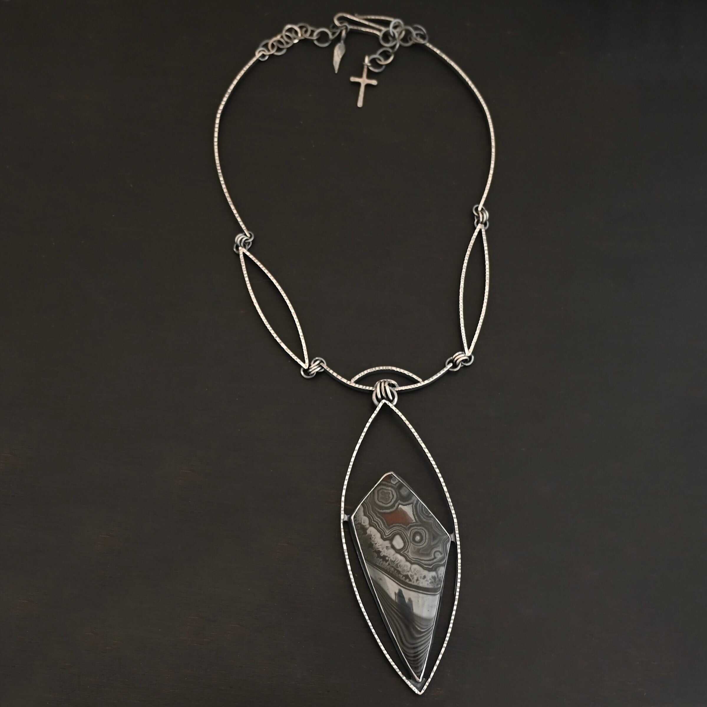 Zinnia Necklace New "Zinnia" Necklace. There is a large, diamond shaped black, grey, and red stone. There is a marquise shape of silver that goes around the stone, connected on each side to the stone. The chain of the necklace has two marquise shapes of silver, one on each side. The center is a small curve of silver from which the jump rings hang from that are attached to the marquise of silver.