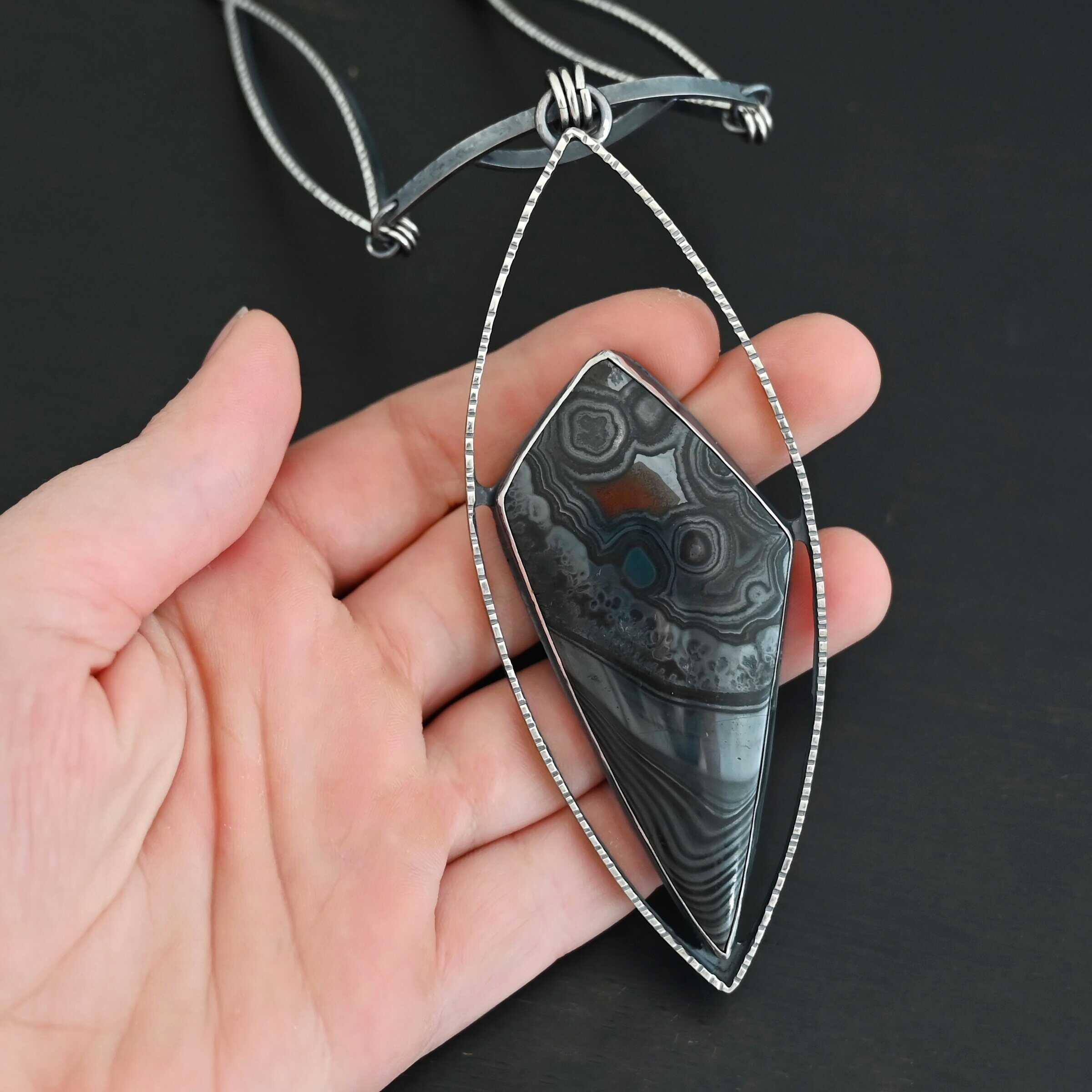 Zinnia Necklace New "Zinnia" Necklace. There is a large, diamond shaped black, grey, and red stone. There is a marquise shape of silver that goes around the stone, connected on each side to the stone. The chain of the necklace has two marquise shapes of silver, one on each side. The center is a small curve of silver from which the jump rings hang from that are attached to the marquise of silver.