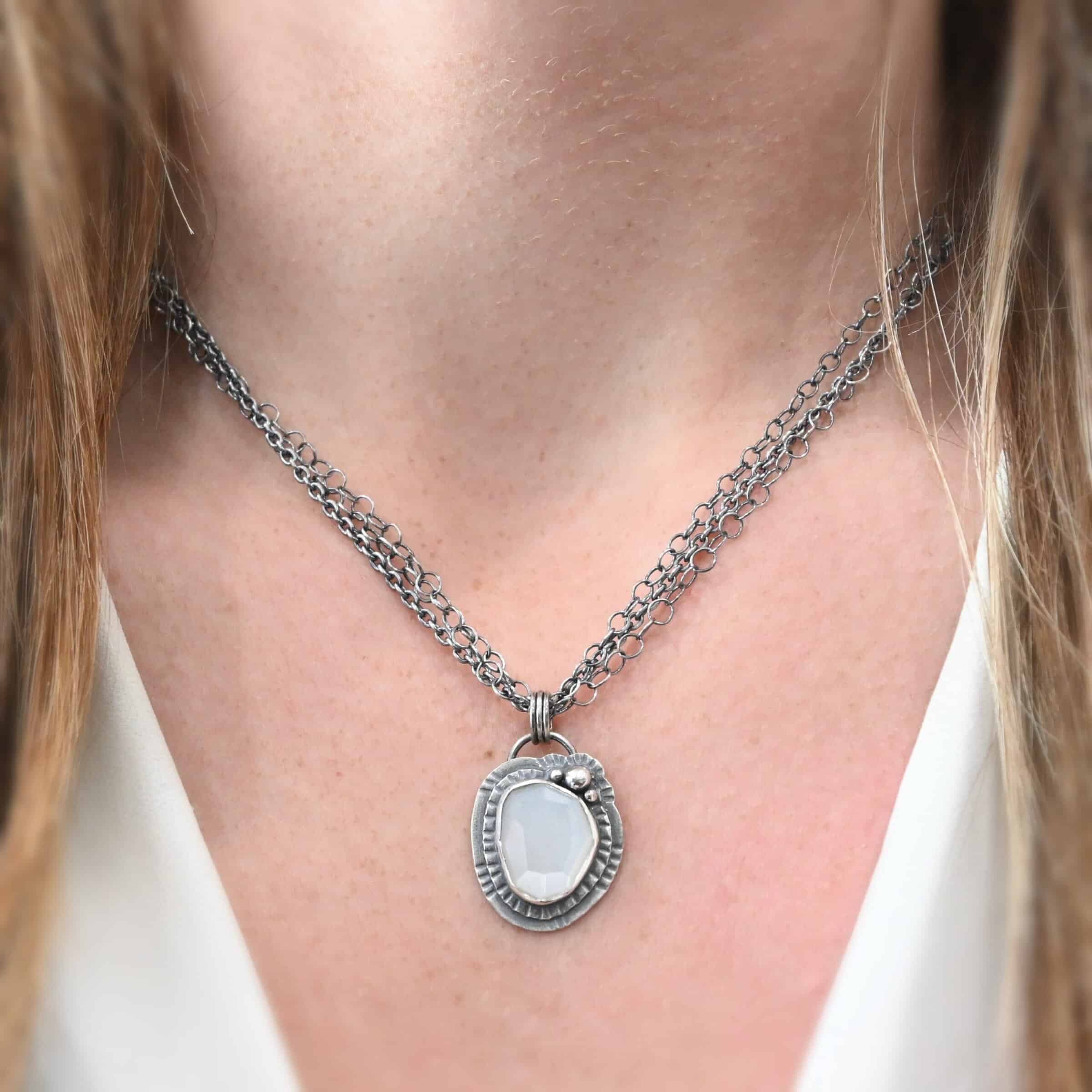 Inara Necklace "Inara" Necklace. There is an oval, vertical moonstone in the center. There are three balls of silver on the top right side. There are two layers of hammered sterling silver around the rest of the stone. The chain of the necklace is multiple chains that meet at the back. Necklace is shown on a model.