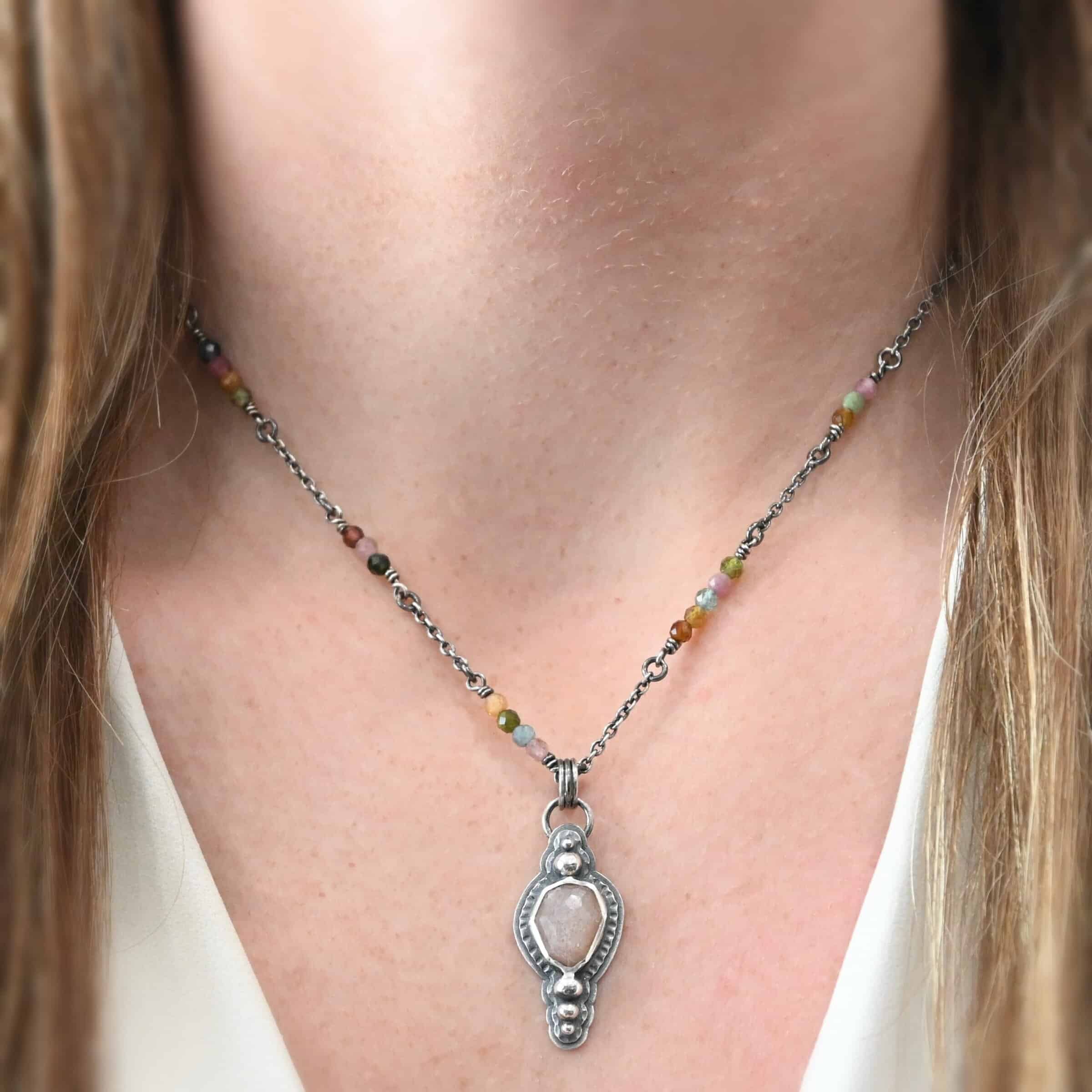 Leo Necklace "Leo" Necklace. There is an oval peach moonstone in the center. There are two balls of silver above it and three below it. The rest of the silver around the stone is hammered. There is a larger layer behind that which is smooth. There is a loop soldered to the back with three jump rings attached to it. The chain goes through them, and the chain has small tourmaline beads. Necklace is shown on a model.