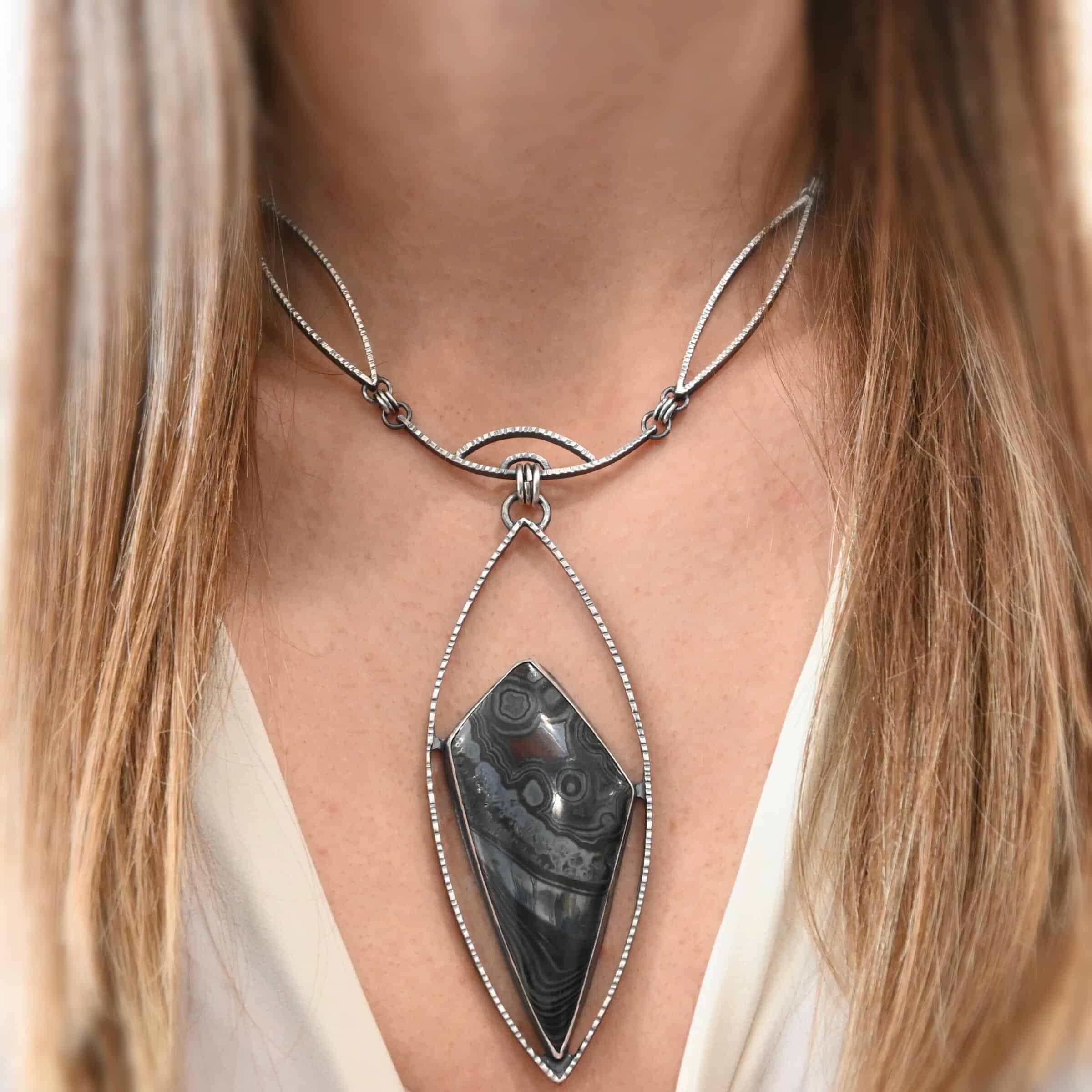 Zinnia Necklace New "Zinnia" Necklace. There is a large, diamond shaped black, grey, and red stone. There is a marquise shape of silver that goes around the stone, connected on each side to the stone. The chain of the necklace has two marquise shapes of silver, one on each side. The center is a small curve of silver from which the jump rings hang from that are attached to the marquise of silver.