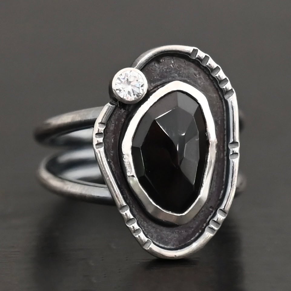Kala Ring Photo of the "Kala" Ring. There is an oval black onyx in the center with a small circular cz above it on the left side. It is surrounded by oxidized sterling silver with a thin line of shiny sterling silver around it with hammered lines in it.
