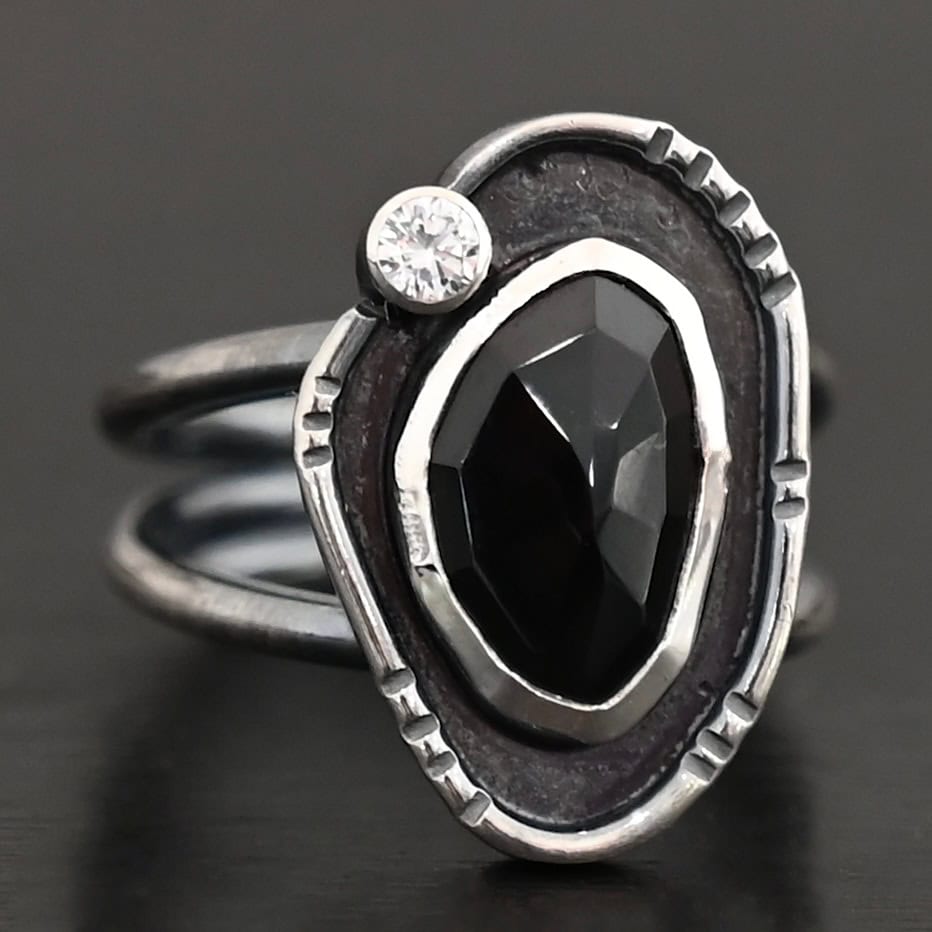 Kala Ring Photo of the "Kala" Ring. There is an oval black onyx in the center with a small circular cz above it on the left side. It is surrounded by oxidized sterling silver with a thin line of shiny sterling silver around it with hammered lines in it.
