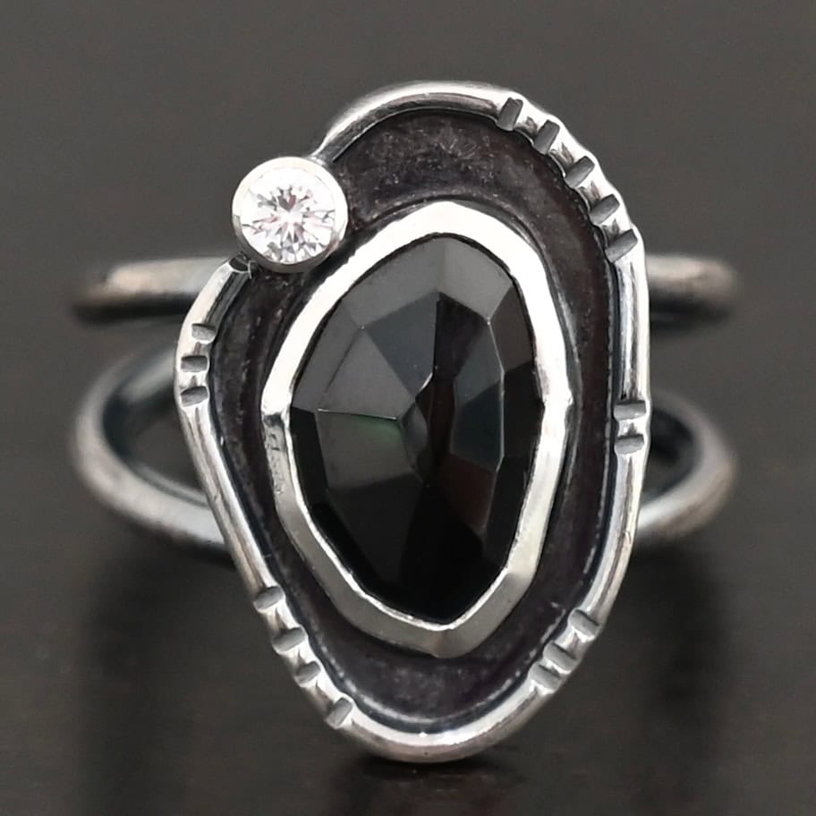 Kala Ring Photo of the "Kala" Ring. There is an oval black onyx in the center with a small circular cz above it on the left side. It is surrounded by oxidized sterling silver with a thin line of shiny sterling silver around it with hammered lines in it.