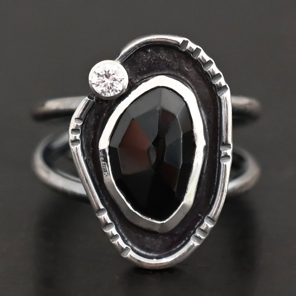 Kala Ring Photo of the "Kala" Ring. There is an oval black onyx in the center with a small circular cz above it on the left side. It is surrounded by oxidized sterling silver with a thin line of shiny sterling silver around it with hammered lines in it.
