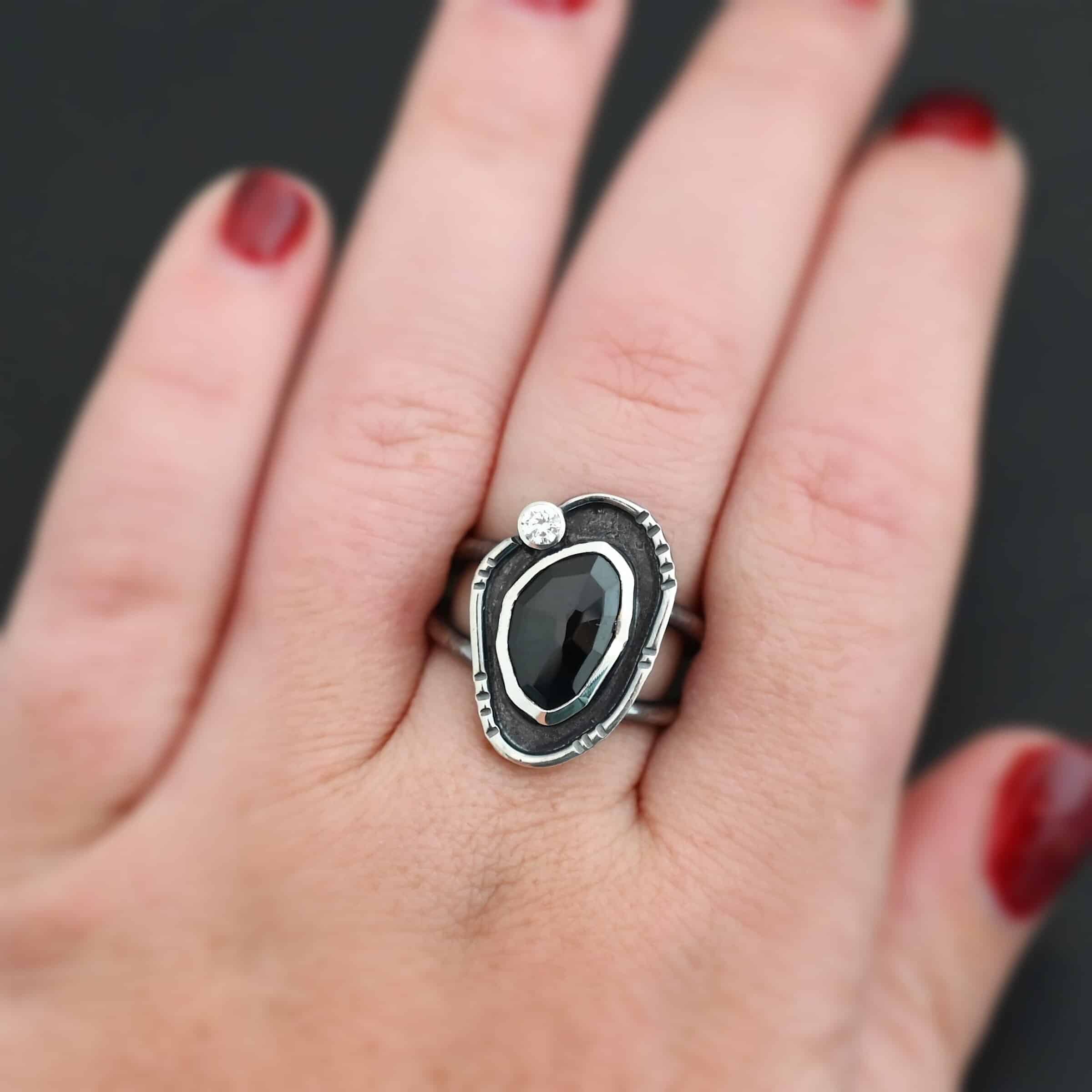 Kala Ring Photo of the "Kala" Ring. There is an oval black onyx in the center with a small circular cz above it on the left side. It is surrounded by oxidized sterling silver with a thin line of shiny sterling silver around it with hammered lines in it. Ring is shown on a hand to demonstrate size.