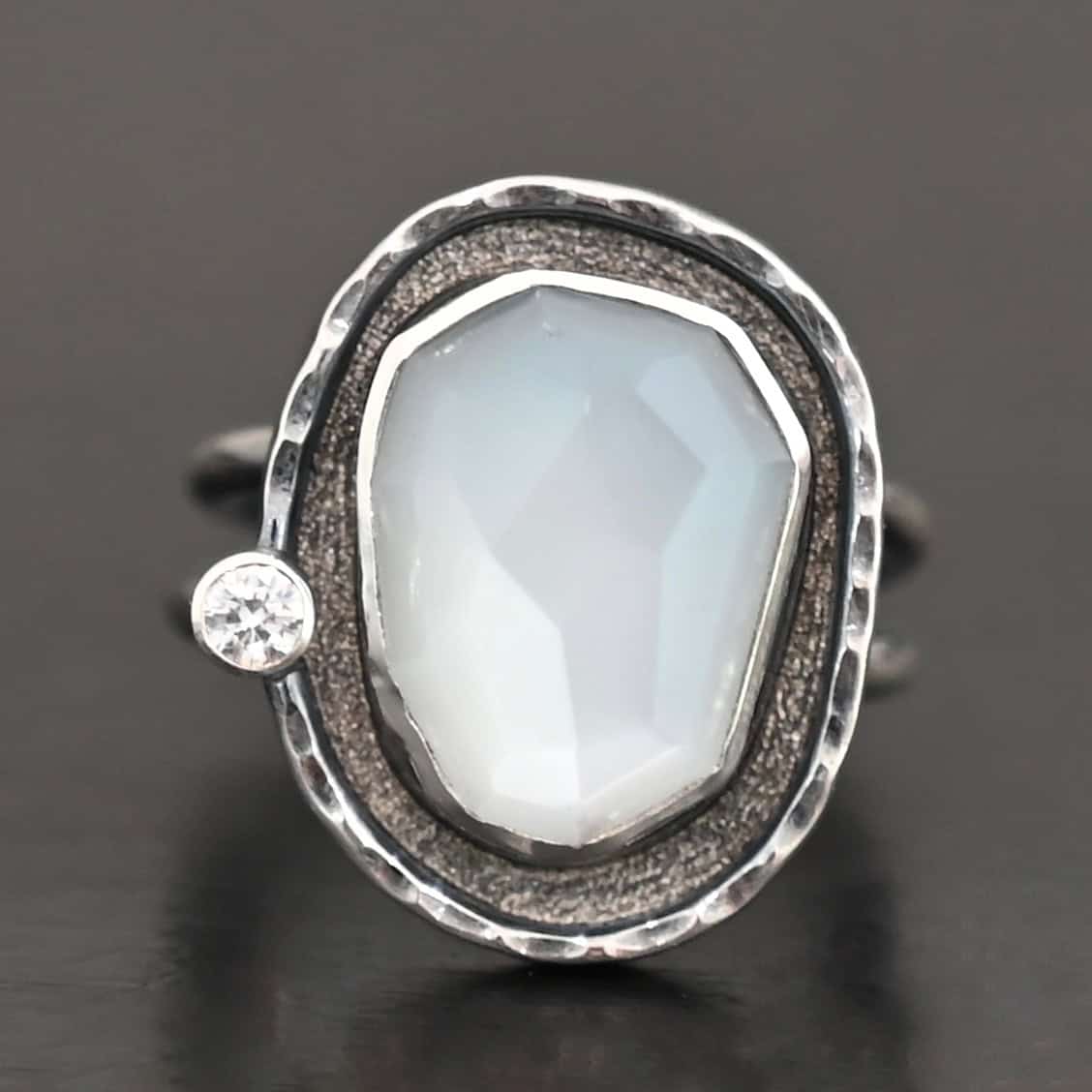 Photo of the "Lainey" Ring. There is an oval moonstone in the center with a small circular cz on the left side. It is surrounded by oxidized sterling silver with a thin line of shiny sterling silver around it that is hammered.