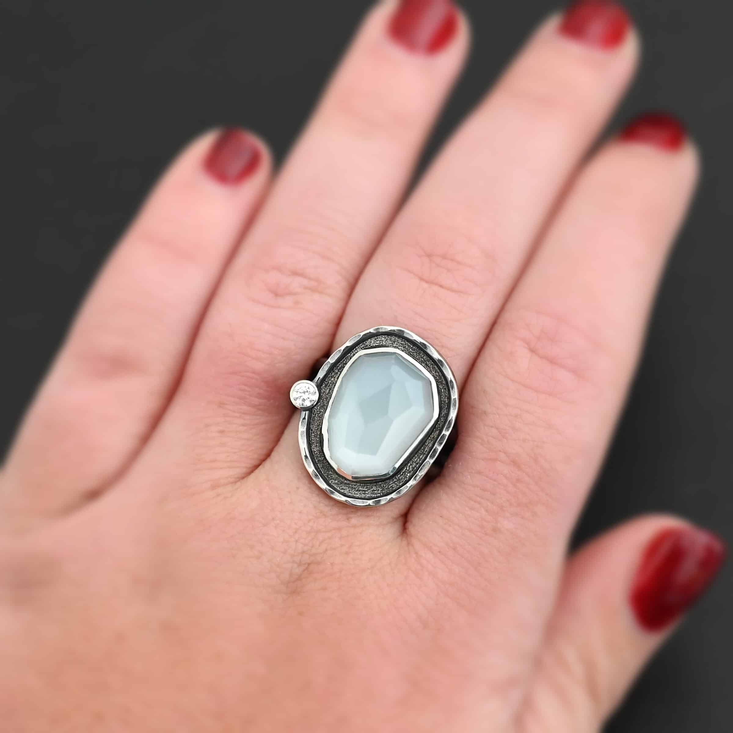 Photo of the "Lainey" Ring. There is an oval moonstone in the center with a small circular cz on the left side. It is surrounded by oxidized sterling silver with a thin line of shiny sterling silver around it that is hammered. Ring is shown on a hand to demonstrate size.