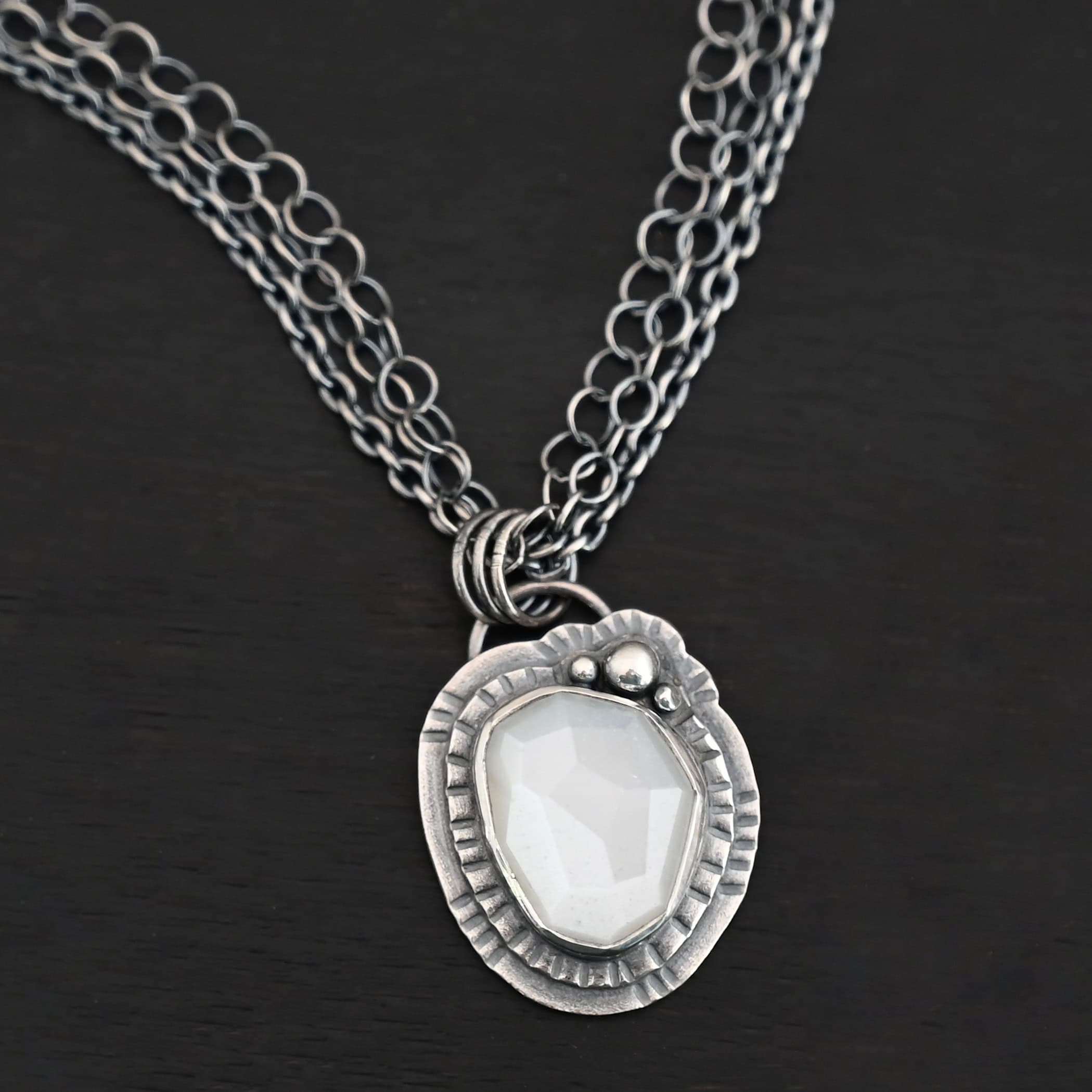 Inara Necklace "Inara" Necklace. There is an oval, vertical moonstone in the center. There are three balls of silver on the top right side. There are two layers of hammered sterling silver around the rest of the stone. The chain of the necklace is multiple chains that meet at the back.